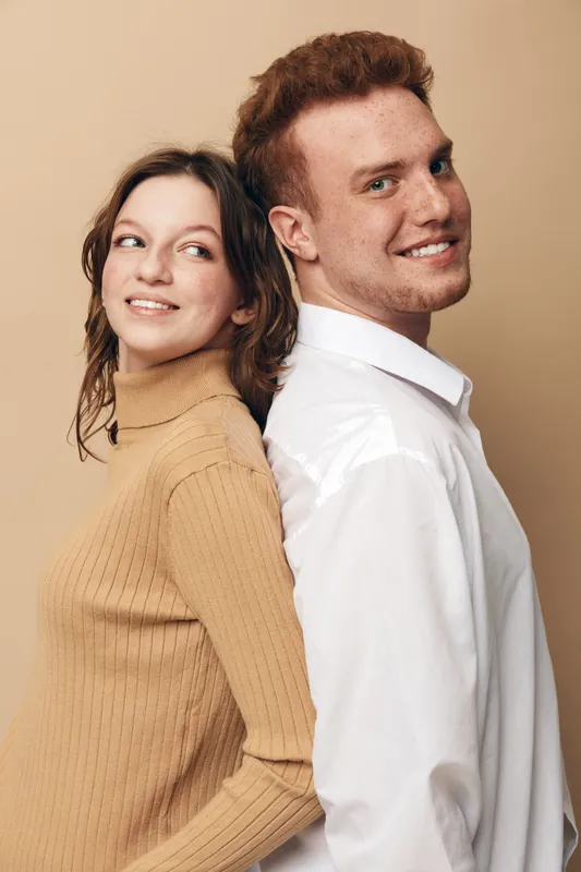 Young man and woman stand back to back smiling, wearing casual clothes on beige background. Concept of friendship, relationship, happiness, and positive emotions.