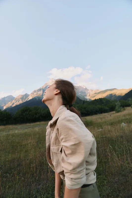 Young woman wearing a beige jacket stands in a grassy meadow looking up toward distant mountains, capturing a moment of calm wonder outdoors and bathed in warm light