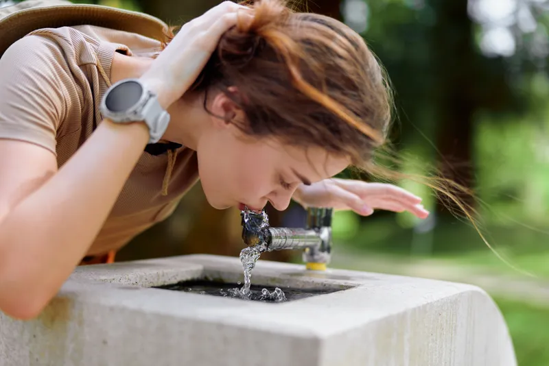 Woman drinking water from a public fountain in a park, leaning forward to cup hands, summer casual outfit with wristwatch visible, natural greenery and outdoor refreshment moment.
