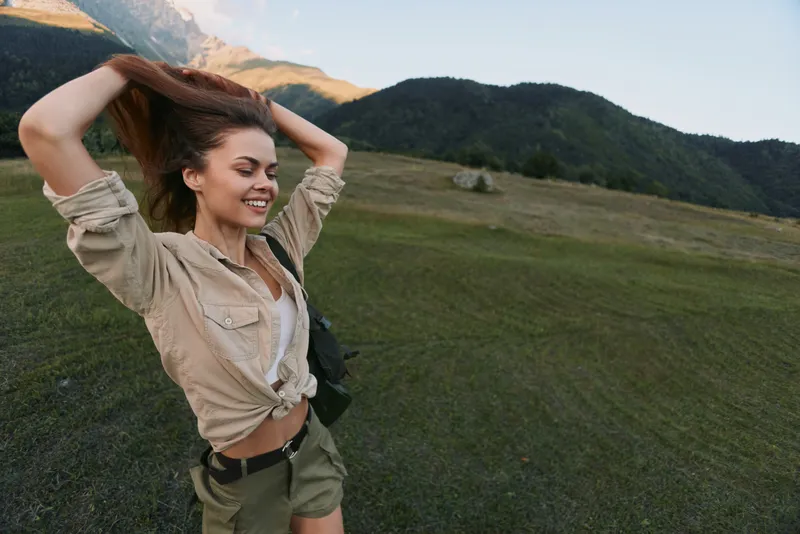 Happy woman with long hair wearing casual clothes enjoys outdoor nature walk in green field near forest and hills, lifestyle concept with fresh air and freedom feeling.