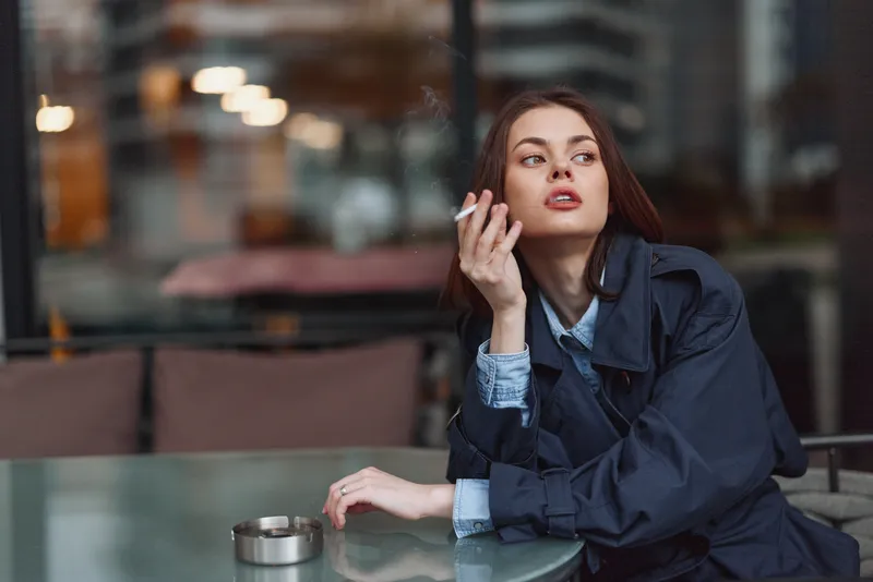 Pensive woman at an outdoor cafe, cigarette in hand, urban lights blurred behind, wearing a trench coat and denim shirt, relaxed pose, contemplative mood, soft evening light