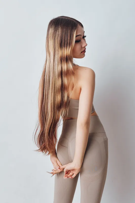 Young woman with long hair in beige outfit posing sideways with hands behind back on light background for fashion beauty and elegance.