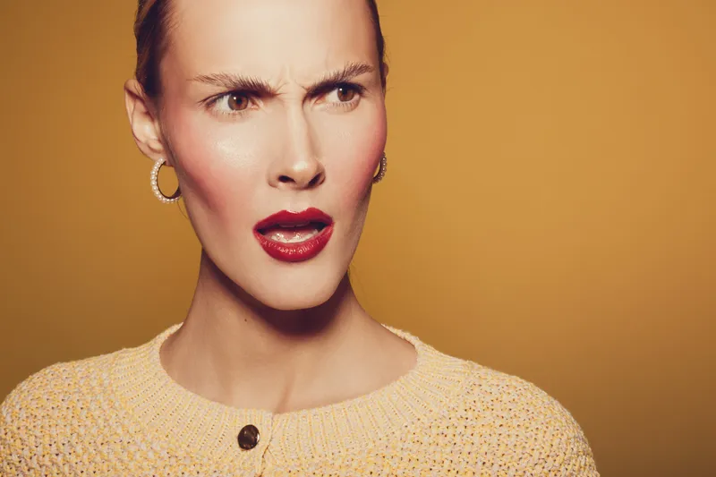 Confused woman with red lipstick and hoop earrings looks surprised against yellow background. Close-up portrait of young female expressing disbelief or shock in casual sweater.