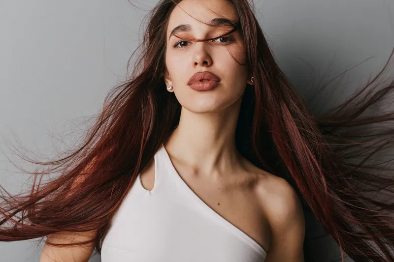 Woman with long brown hair and natural makeup wearing white top looking confidently at camera with hair flowing against gray background in studio portrait.