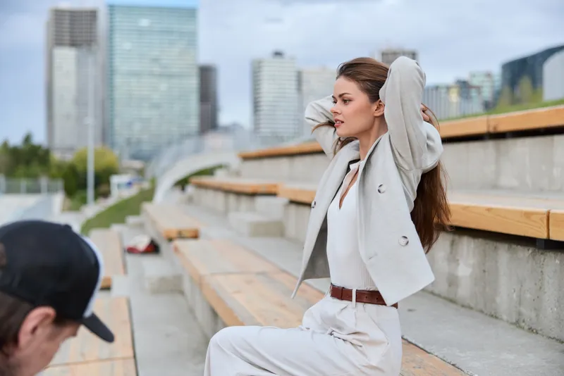 young woman sitting on urban wooden steps outdoors in business casual clothes adjusting hair with city skyscrapers in background