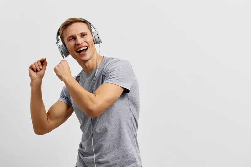 Smiling young man wearing headphones dancing joyfully against a plain white background, expressing happiness and energy. Studio portrait capturing carefree lifestyle, music enjoyment, and positive emo