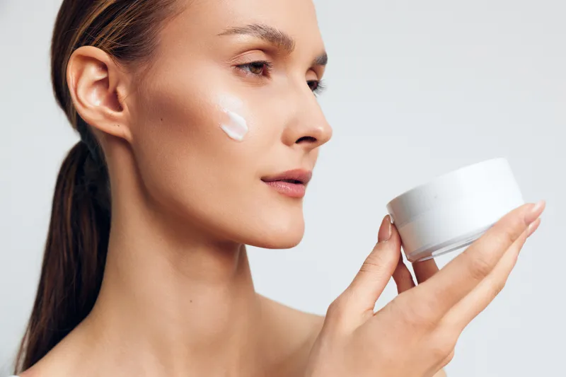Close up of a young woman with brunette hair applying moisturizer to her cheek, showcasing a calm skincare moment with a clean minimalist look and soft studio lighting.
