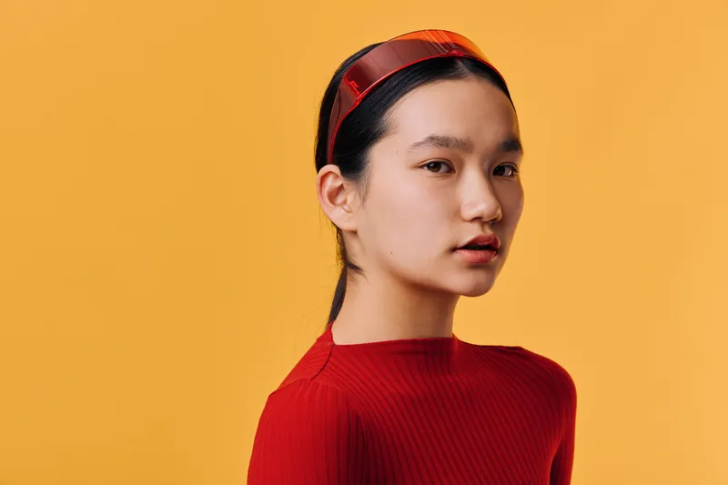 Portrait of young woman in red sweater and headband looking at camera with serious expression on orange background in studio lighting for fashion or beauty concept.