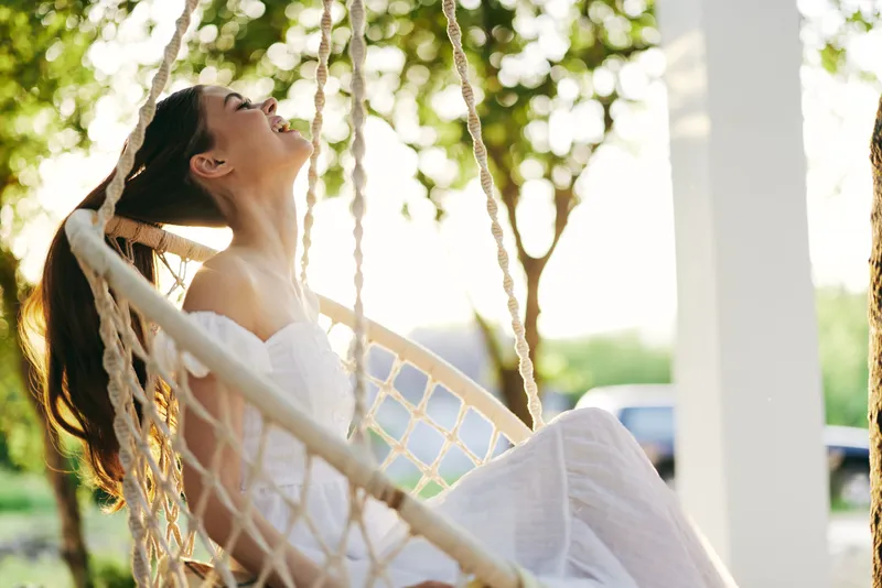 Young woman in white dress enjoying happiness on a swinging chair outdoors, with a sunny background and greenery reflecting joy, beauty, and relaxation