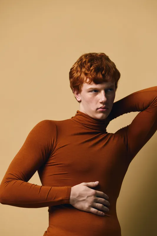 Young man with red hair wearing brown turtleneck posing with hand on head and stomach against beige background, fashion model portrait with serious expression.