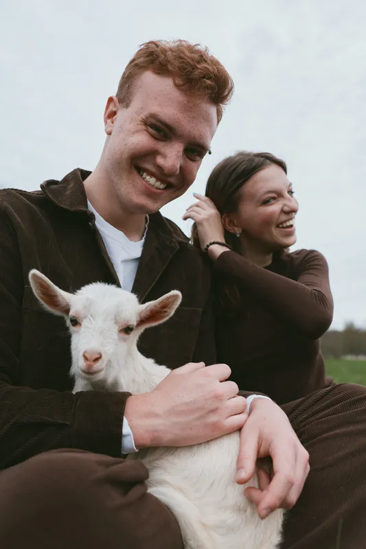 Couple goat kid outdoors happiness lifestyle countryside portrait affection smiling, a candid scene of a joyful pair with a white goat kid in a rural field, warm light, relaxed mood, natural