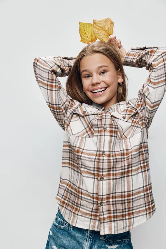 Girl smiling and posing with autumn leaves on head wearing plaid shirt and jeans against white background. Happy child having fun with fall season nature concept.
