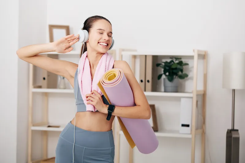 Active woman in a bright home gym enjoying a workout, wearing wireless headphones, carrying a yoga mat and towel after a session, with shelves, plant and minimalist decor in the background.