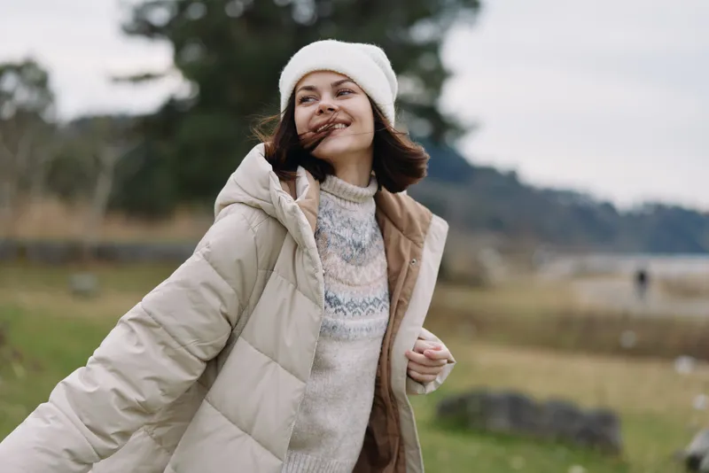 Woman in a warm coat and knit sweater enjoying a breezy outdoor setting, smiling, wearing a beanie, with a blurred countryside background.