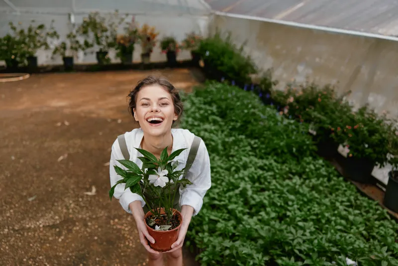 Young woman joyfully holding a potted plant in a greenhouse, showcasing vibrant greenery and a sense of connection with nature for a healthy lifestyle