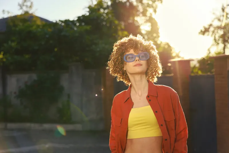 A stylish young woman with curly hair stands outdoors in bright sunlight, wearing a yellow crop top and red jacket, modeling bold streetwear for a lifestyle shoot