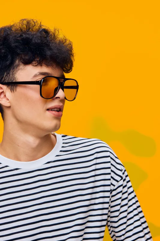 young man wearing striped shirt and stylish sunglasses with curly hair looking sideways against bright yellow background in casual fashion style