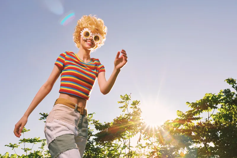 Cheerful woman outdoors, smiling under a bright sun, striped top and casual shorts, relaxed pose, warm breeze, vibrant colors, natural greenery and clear sky in the background through trees