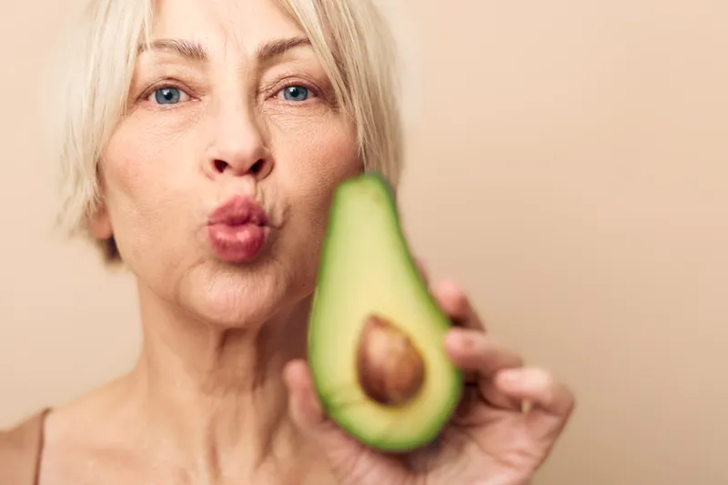 Mature woman with short blonde hair holds half of avocado close to camera while puckering lips. Natural skin and healthy food concept with fresh fruit and positive expression.
