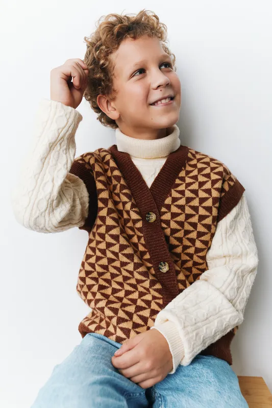 Young boy wearing a cozy brown and cream geometric knit vest sits gently against a white wall, smiling softly with a relaxed, thoughtful pose and warm casual mood.