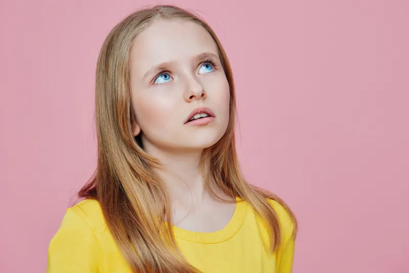 Young girl with long blonde hair looking up against a pink background, wearing a yellow shirt, expressing curiosity and innocence Perfect for children s themes