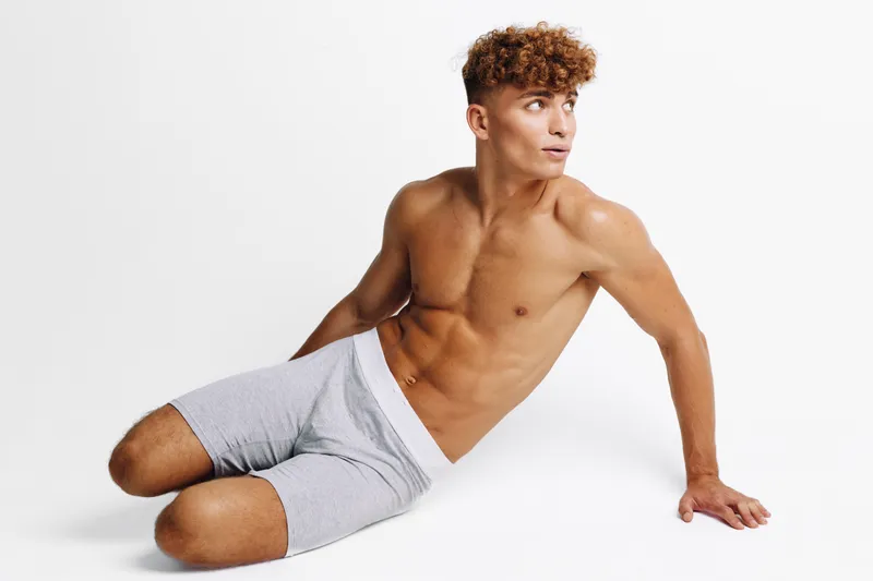 Young man with muscular body sitting on floor in casual shorts looking sideways with thoughtful expression on white background. Fitness, health and relaxed pose for lifestyle concept.