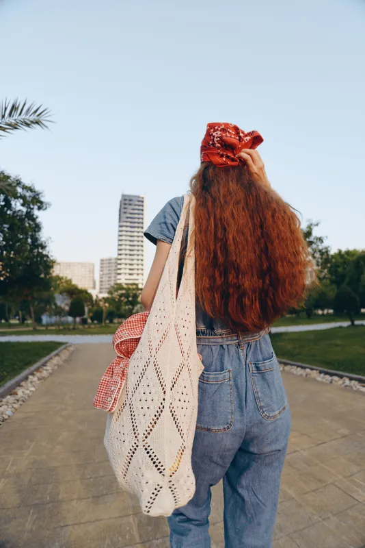 lifestyle woman with long hair wearing modern boho-western denim street style and red bandana walking outdoors with crochet bag in film color tone