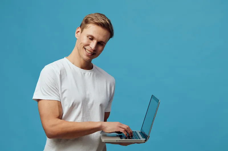 Smiling young man in casual white t-shirt using a laptop computer isolated on a plain blue background. Studio portrait showing technology, work, and casual lifestyle concepts.