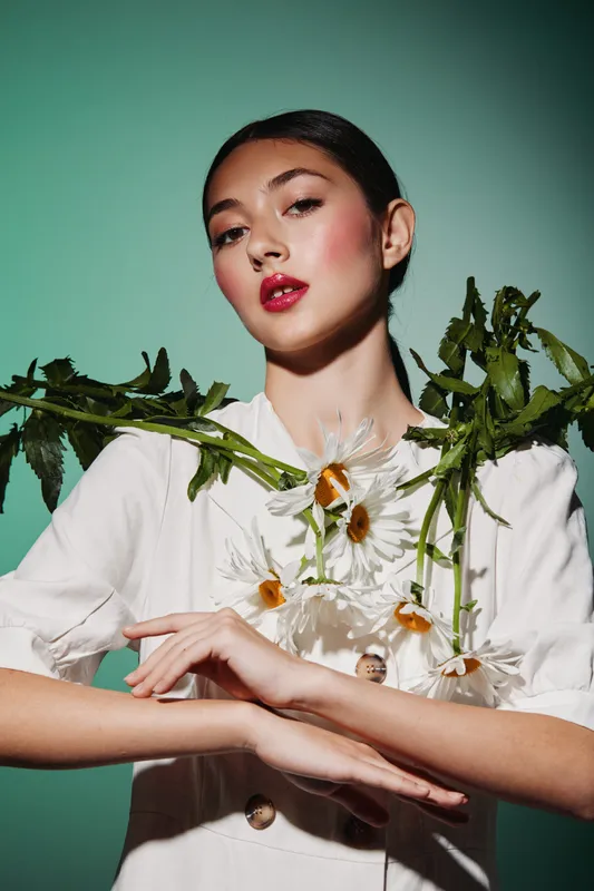 Stylish young woman with glossy red lips and rosy blush poses with wildflowers on shoulders against a turquoise background. Creative beauty and fashion concept showing elegance.