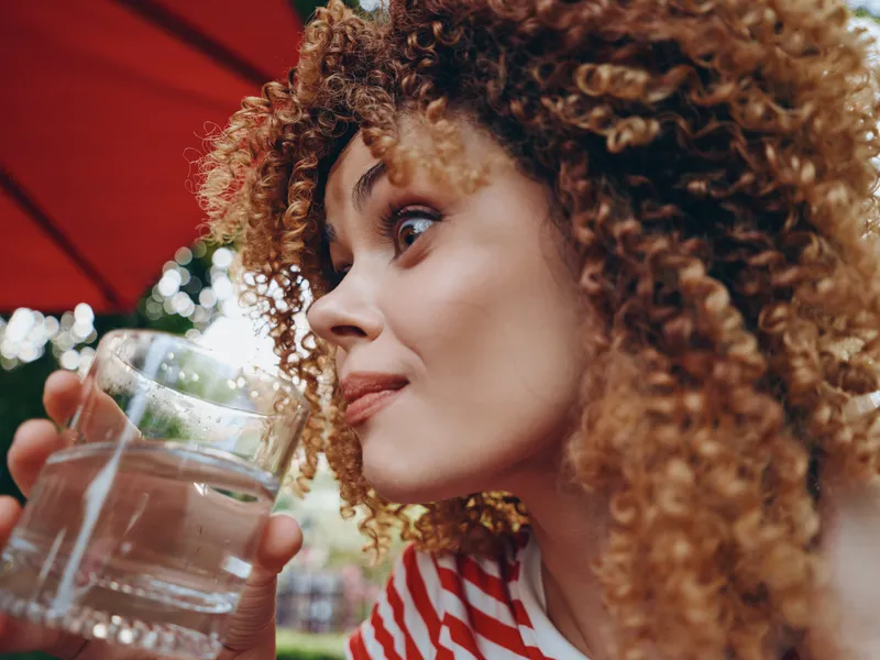 Close-up of a joyful woman with curly hair sipping water from a clear glass outdoors under a red umbrella, capturing a fresh summer mood and candid expression.