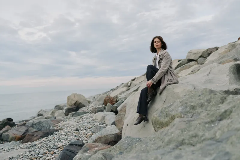Young woman with dark hair wearing casual clothes sits on large rocks by the calm sea under cloudy sky. Concept of solitude, contemplation, and nature connection in outdoor setting.