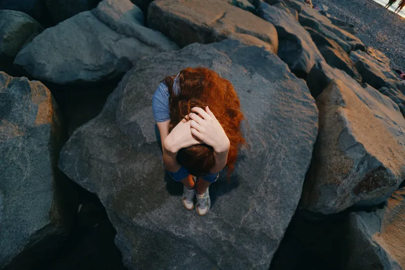 lifestyle woman with red hair in modern boho-western denim style sitting on rocks, street fashion vibe, film color tones, casual outdoor relaxation mood