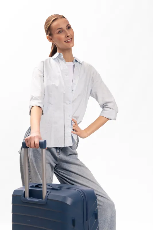 Young woman dressed in casual clothes stands with hand on hip holding suitcase handle, ready for travel. Concept of vacation, business trip, and lifestyle with luggage on white background.