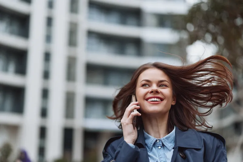 Young woman with long brown hair smiles happily while talking on a smartphone outdoors in an urban environment with modern buildings behind her.