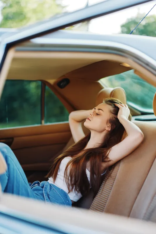 Young woman relaxing in a car with long hair, wearing casual attire, enjoying a carefree moment in nature, showcasing a serene expression and vibrant colors.