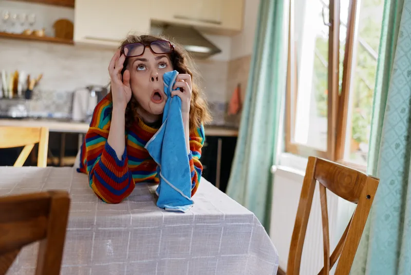 A stressed woman sits at a kitchen table in a bright striped sweater, head resting on a cloth, phone in hand. The scene conveys overwhelm, fatigue, and mental strain in a domestic setting.