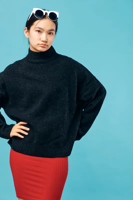 Young woman posing in black sweater and red skirt against blue background with white sunglasses on head, looking confident and casual with hand on hip in fashion portrait style.