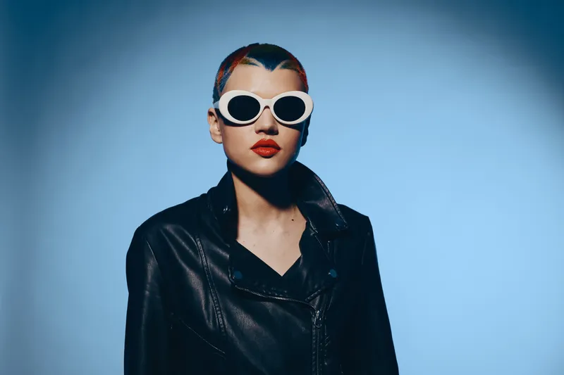 Young woman with short hair wearing white sunglasses and black leather jacket standing against blue background, stylish and bold contemporary fashion portrait.