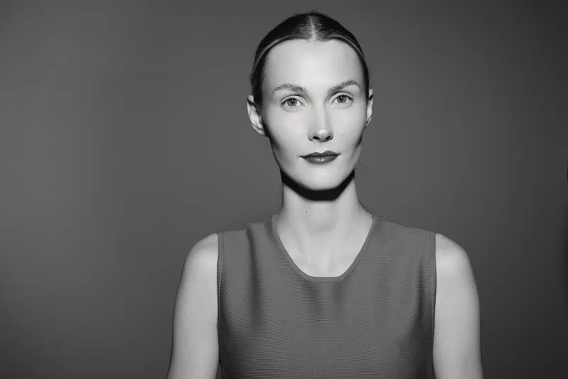 Portrait of a caucasian woman with fair skin and tied hair wearing a sleeveless top. The black and white studio photo features a neutral expression and simple background.