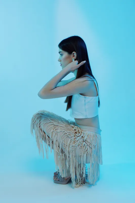 Young woman in white top and fringed pants poses in profile against blue background. Concept of beauty, fashion, and modern style with soft studio lighting.