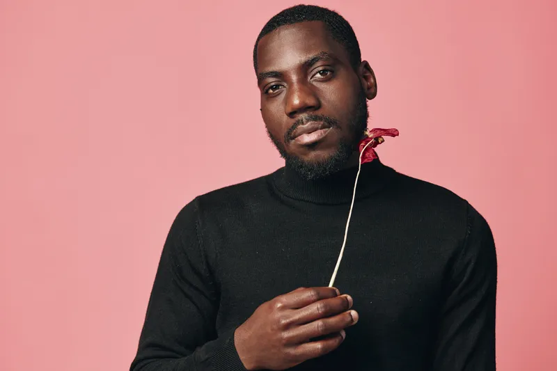 African man with dark skin holding red flower in creative conceptual visual style studio portrait. Black sweater, pink background, thoughtful expression, isolated, artistic and modern photography.