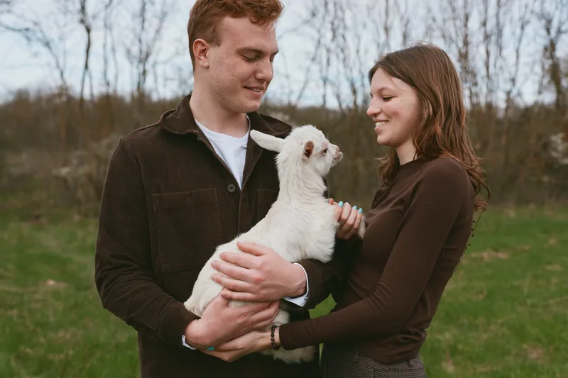 A couple outdoors holds a white goat kid, sharing a warm affectionate moment in a grassy countryside, smiling faces and rustic setting, calm, loving interaction with farm animal