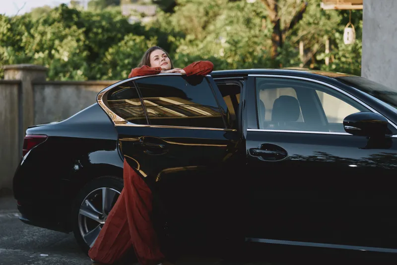 Cheerful woman in a red dress leans against a sleek car outdoors, sunlit moment, relaxed pose, natural colors, candid lifestyle scene
