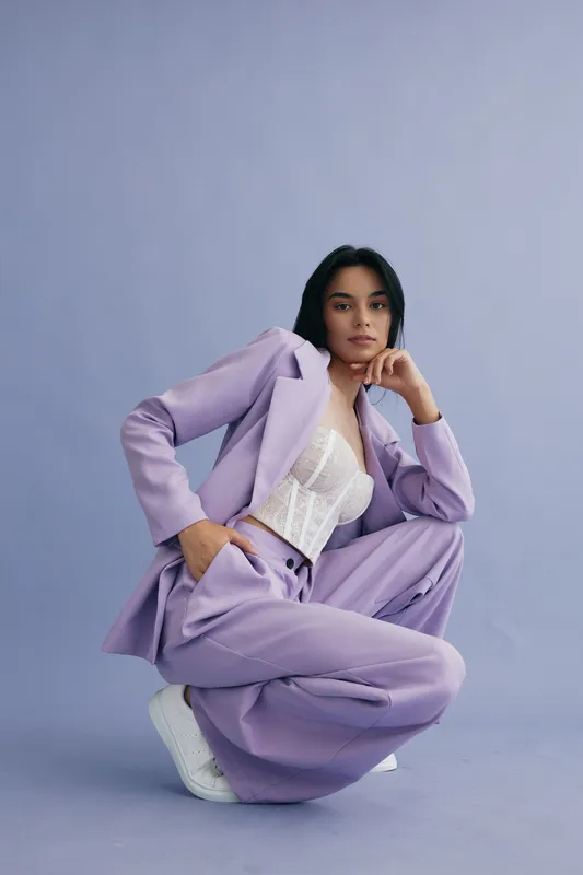 Stylish woman in a lilac pantsuit and white lace top posing confidently against a soft purple background, showcasing modern fashion and casual elegance.