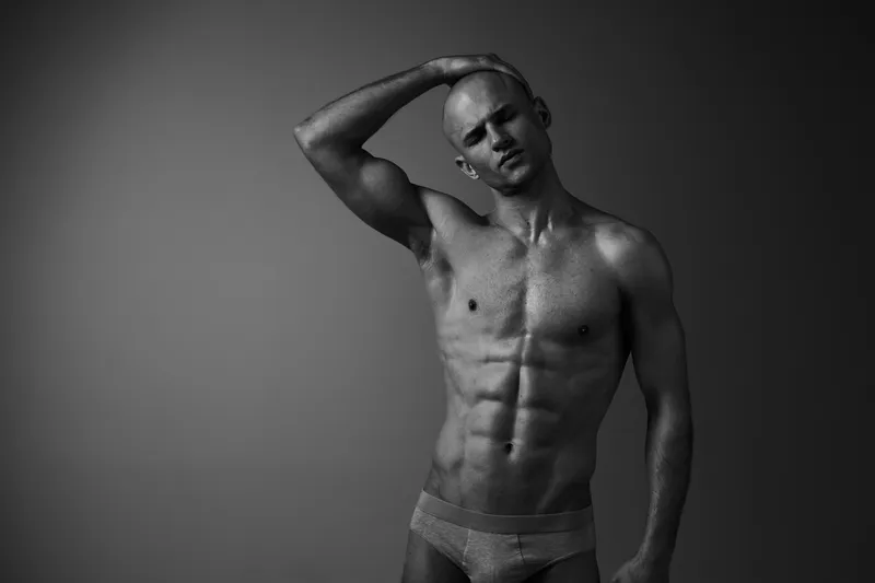 Black and white portrait of a man posing confidently, showcasing his toned physique and captivating expression. A striking image that emphasizes strength and elegance.