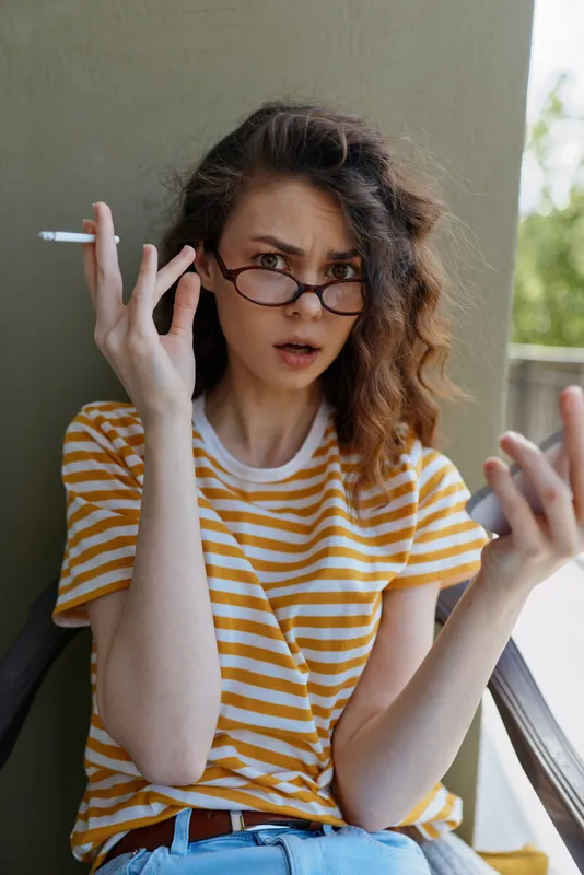 A surprised woman sits on a balcony wearing a striped yellow shirt and glasses, cigarette in hand, mid gesture showing confusion. Concept for startled mood and casual home life.