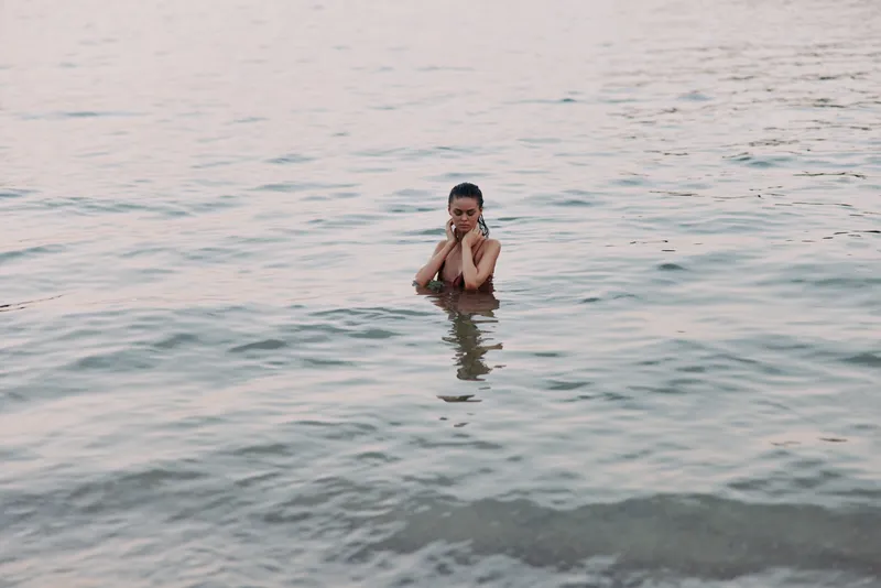 A solitary figure stands waist deep in calm sea water, hands near face, gentle ripples and soft light create a tranquil, contemplative moment in a serene seascape.