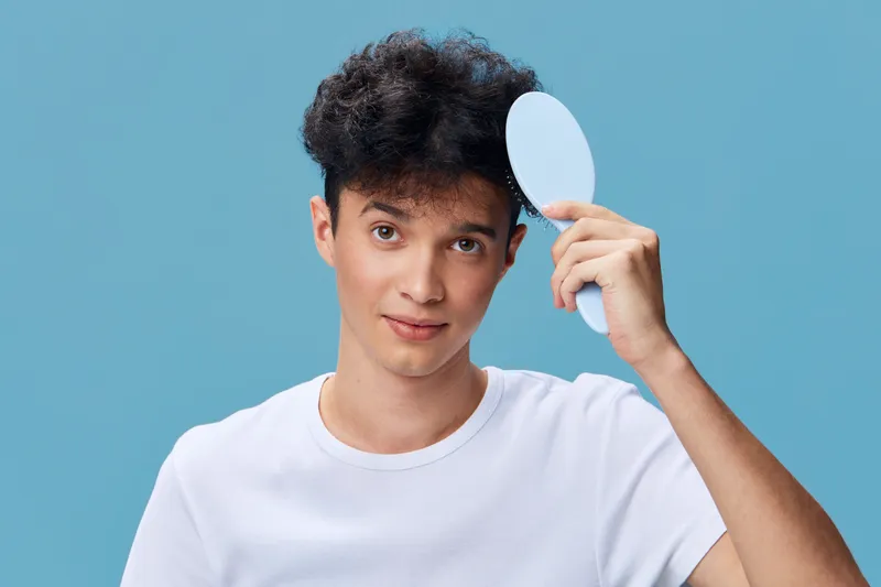 Young man with curly hair holding a round mirror near his face, wearing a white t-shirt on a blue background, looking curious and thoughtful.
