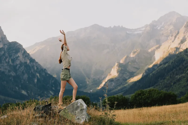 Young woman stands on a rocky outcrop in a mountain meadow, arms raised in triumph and freedom