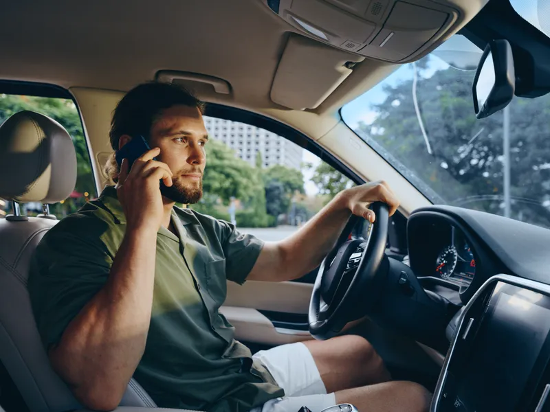 Portrait of a man driving a car while talking on a smartphone, calm expression, casual shirt, visible steering wheel, dashboard, and interior detail in a modern vehicle.